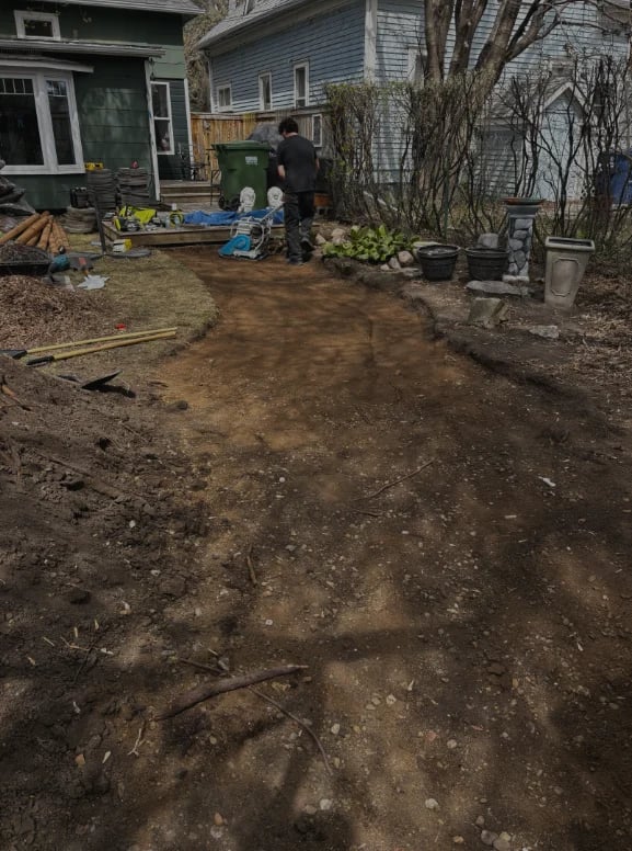 Person working in muddy backyard garden area next to residential house with bare trees and gardening materials