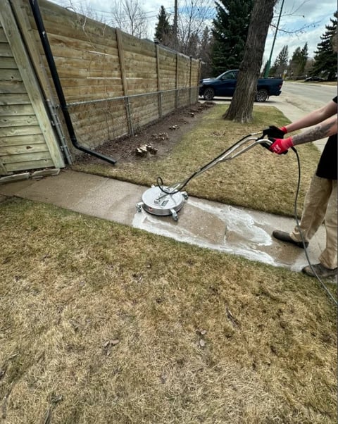 Person using a pressure washer to clean a concrete patio in a residential backyard with wooden fence and trees