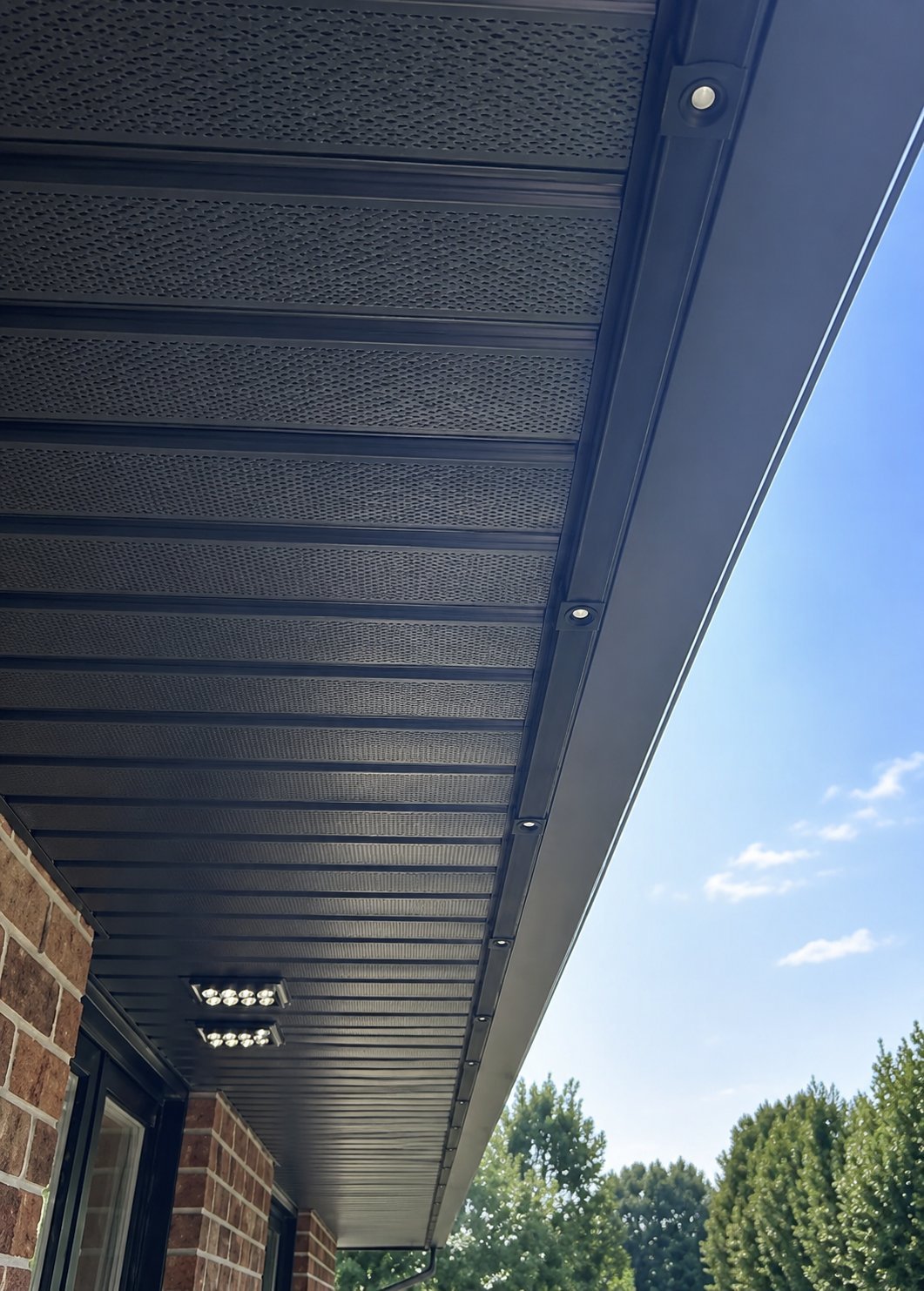 Dark metal pergola or patio cover attached to brick building with integrated LED lights and clear sky visible in background