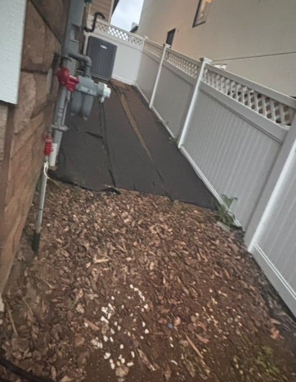 Narrow outdoor passageway with white metal fence, dark gate, brick wall, and gravel ground leading to residential area