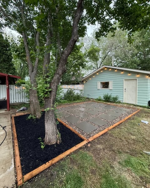 Backyard with light blue shed, large tree, mulched planting area with copper border, and paved patio space
