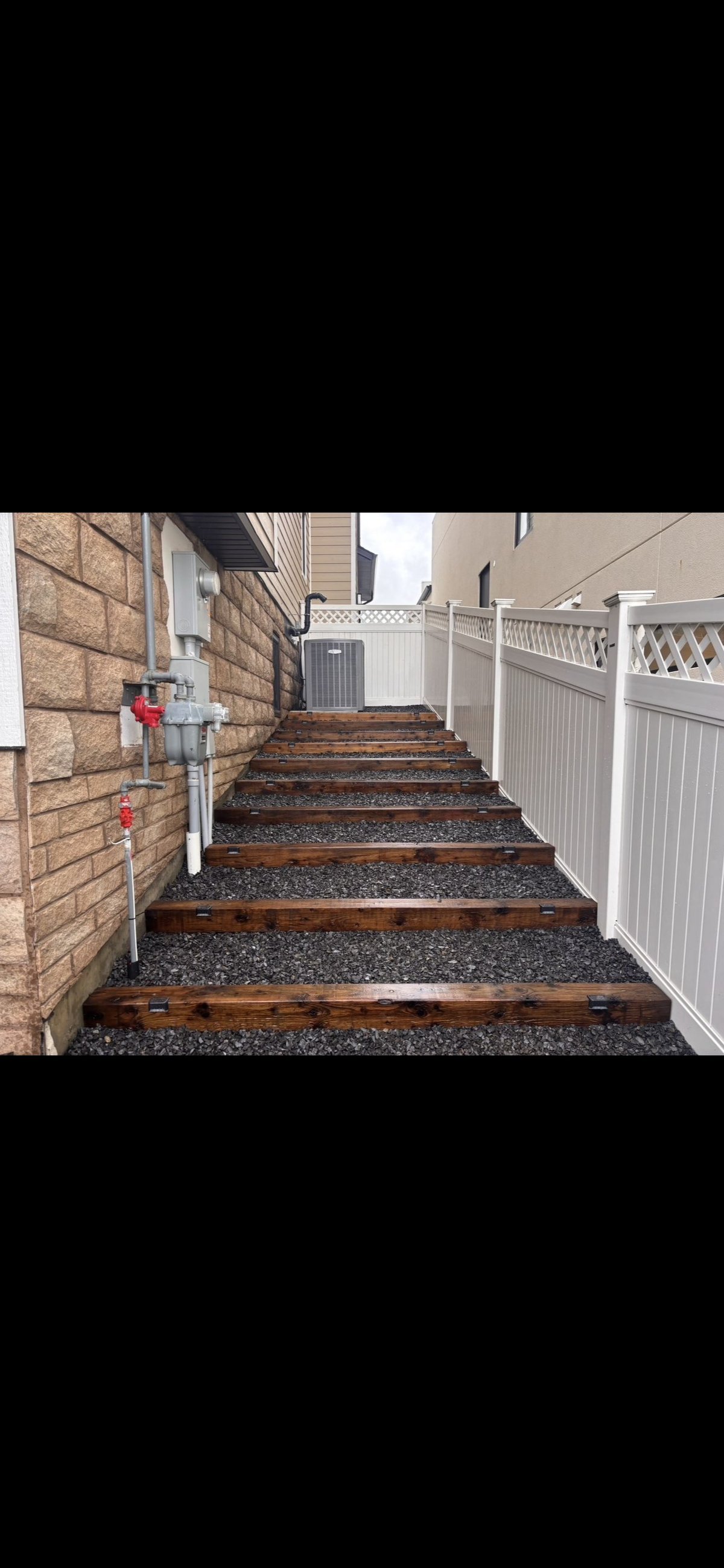 Outdoor stairway with dark gravel treads and wooden risers, flanked by brick wall and white vinyl fencing, leading uphill between residential buildings